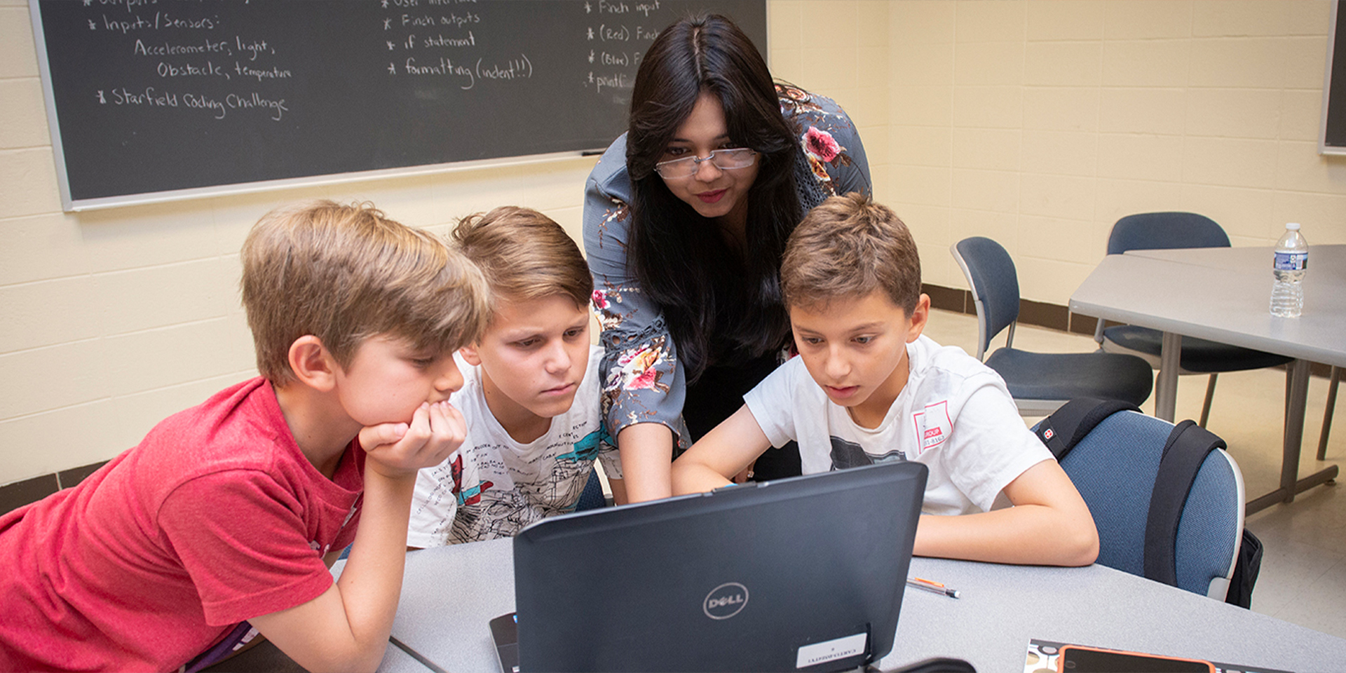 Teacher points to laptop screen as she helps three students with an assignment
